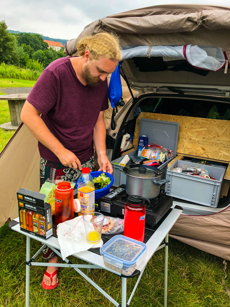 Julian kocht neben unserem Mini-Camper Astrarix. Küche im Camper ist zu sehen sowie das Vorzelt, welches über dem Kofferraum befestigt wird.