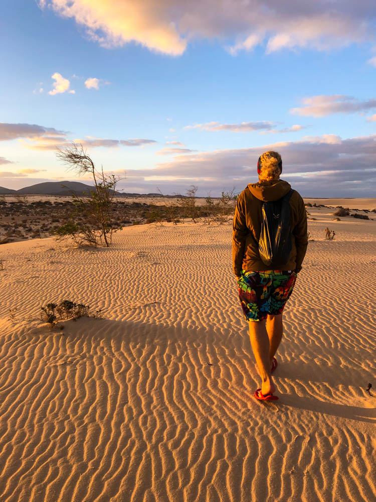 Julian geht in den Dünen vor Corralejo spazieren. Die Sonne geht gerade unter und die wenigen Wolken am Himmel sind leicht orange verfärbt. Erlebnisse auf Reisen