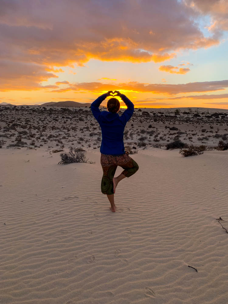Melanie macht eine Yoga Pose in den Sanddünen vor Corralejo. Sie blickt in Richtung des Sonnenuntergangs und formt mit ihren Händen dabei ein Herz. Der Himmel ist komplett orange verfärbt.