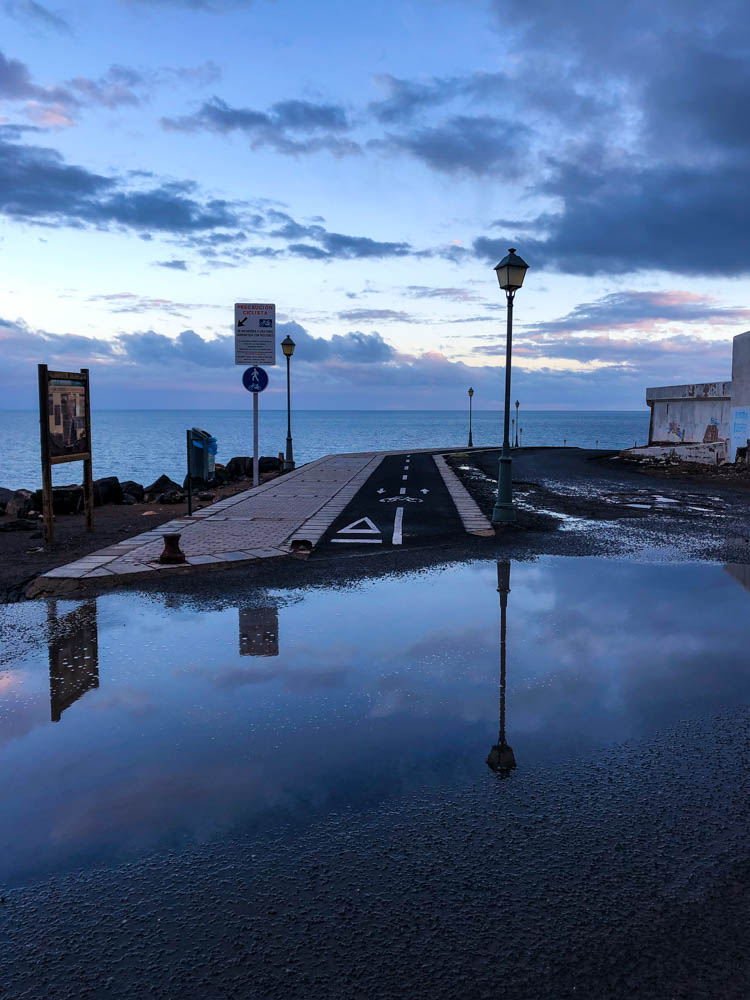 Es ist eine überschwemmte Straße nach einem Regenschauer auf Fuerteventura zu sehen. Der Himmel sowie der im Vordergrund zu sehende Gehweg Abschnitt (inkl. Laterne, Schilder...) spiegeln sich in der Pfütze.