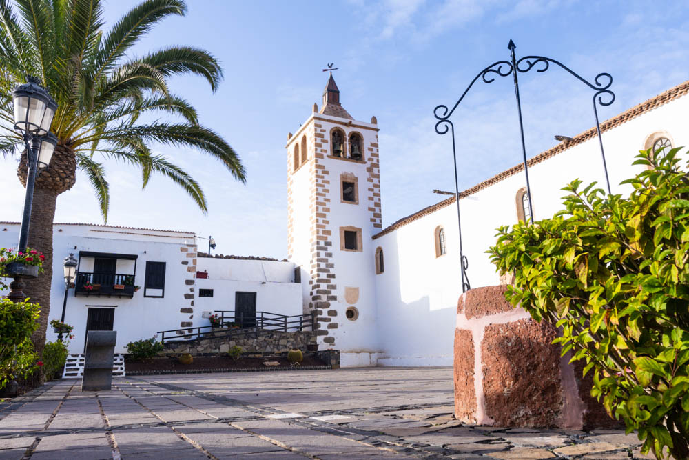 Kirche mit großem Kirchtum in Betancuria. Links im Bild steht eine Palme, rechts vorne eine Brunnen.