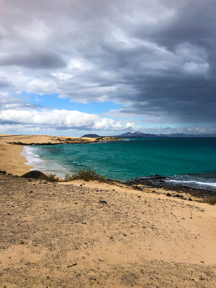 Strand bei den Dünen von Corralejo. Über dem Atlantik, rechts im Bild, hängt eine schwarze Wolkenfront. Das Wasser ist durch den hellen Untergrund türkisblau gefärbt.