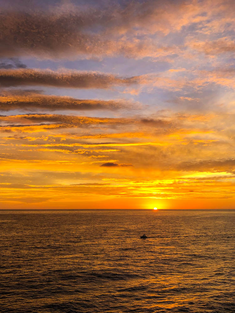 Sonnenaufgang in Fuerteventura über dem Atlantik. Der Himmel sowie das Meer glühen orangefarben. Es fährt gerade ein Boot durch das Bild und die Wolken wechseln die Farbe von orange zu dunkleren Tönen ab.