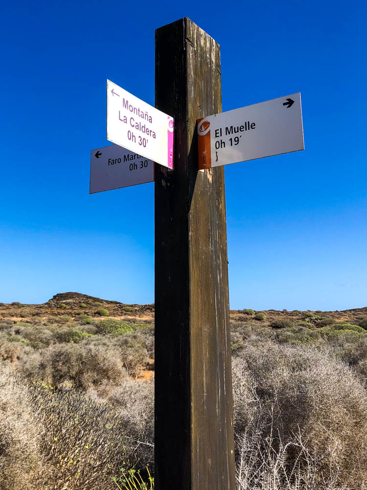 Wegweiser auf der Insel Lobos welcher drei verschiedene Wege beschreibt. Im Hintergrund ganz in der Ferne ist die höchste Erhebung von Lobos zu erkennen.