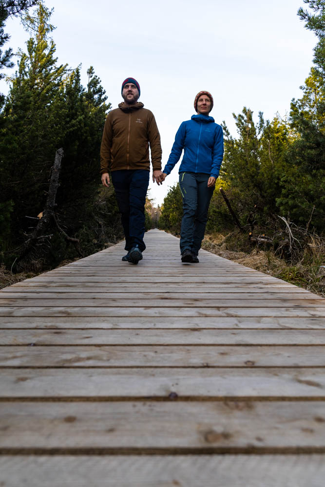 Melanie und Julian laufen Hand in Hand auf einem Holzsteeg auf die Kamera zu. Sie schauen dabei in die Ferne über die Kamera hinweg.