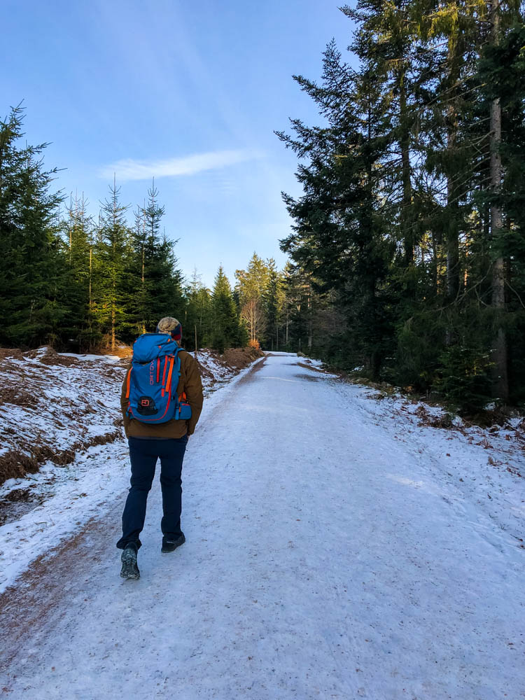 Julian läuft über einen verschneiten Wanderweg. Die Hände hat er in den Hosentaschen. Sein türkiser Rucksack bildet einen Kontrast zu der Waldlandschaft.