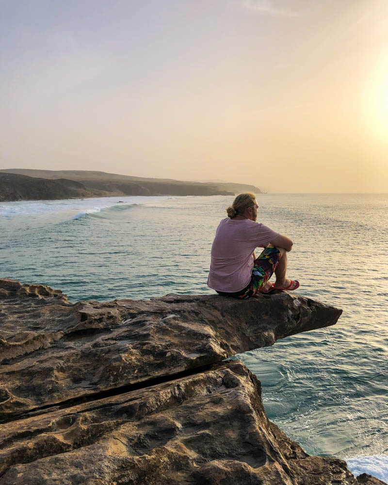 Julian sitzt auf einer Felsenzunge bei La Pared. Er schaut in die Ferne Richtung Sonnenuntergang und beobachtet die Wellen des Atlantiks. Unsere Zeit auf Fuerteventura geht zu Ende