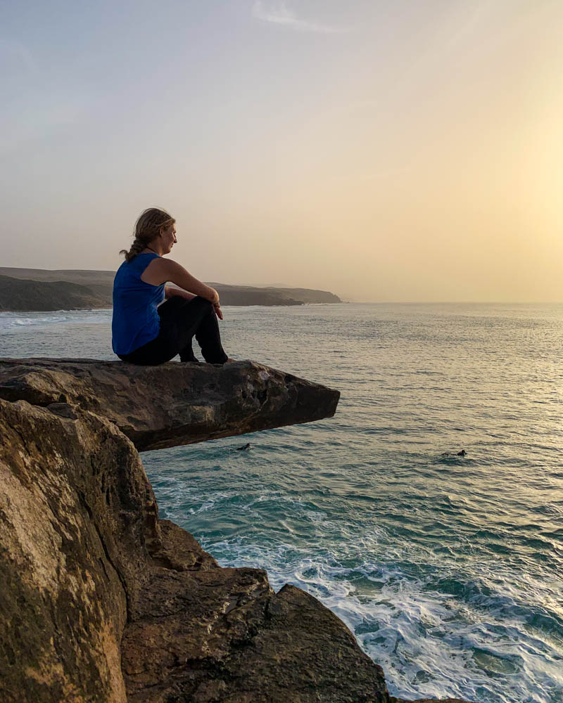 Melanie sitzt auf einer Felsenzunge bei La Pared und schaut den Surfern auf dem Atlantik zu. Der Himmel ist durch den Calima leicht braun verfärbt und die Sonne geht gerade unter. Unsere Zeit auf Fuerteventura geht zu Ende, La Pared zum Abschied