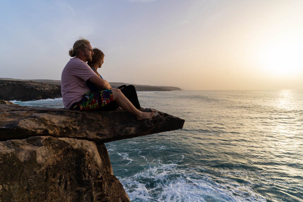 Melanie und Julian sitzen auf einer Felsenzunge und blicken in die Ferne. Die Sonne geht gerade über dem Atlantik unter.