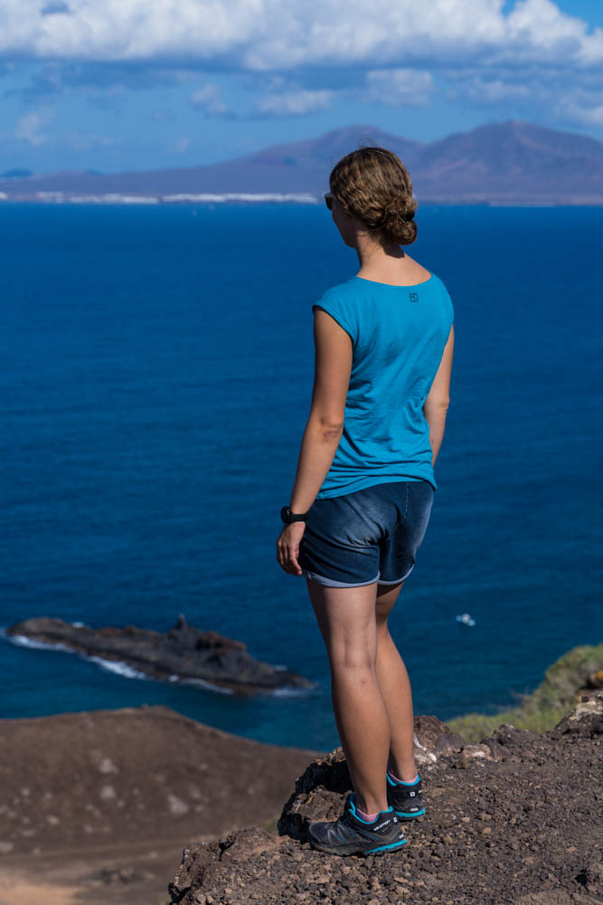 Melanie steht an der Steilküste des höchsten Berges auf Lobos und schaut in die Ferne. Im Hintergrund ist die Insel Lanzarote zu sehen.