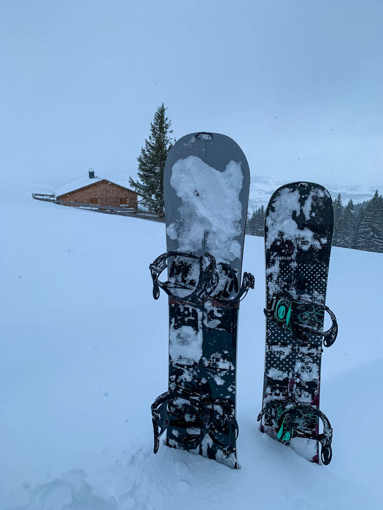 Die zwei zusammengebauten Splitboards stecken im Schnee. Im Hintergrund ist eine Hütte zu erkennen. Der Himmel ist vor lauter Schneefall komplett weiß.