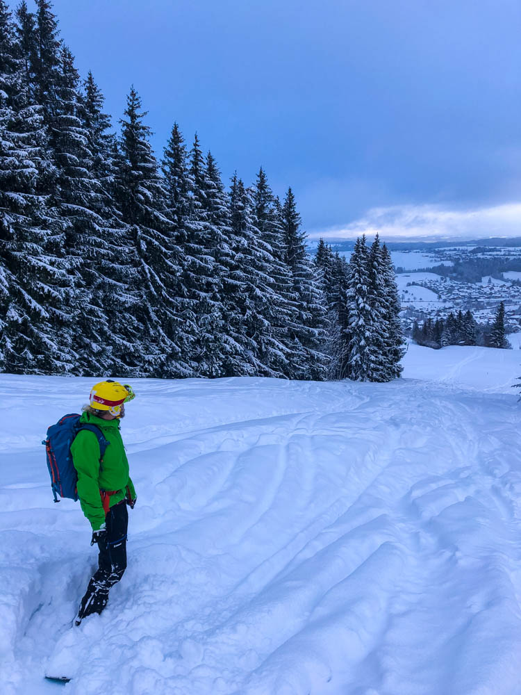 Julian steckt mit dem Snowboard im tiefen Schnee auf der Abfahrt in Nesselwang fest. Sein gelber Helm und die grüne Jacke bilden einen starken Kontrast zur weißen Winterlandschaft. Erste Abfahrt 2021 zurück in Deutschland.