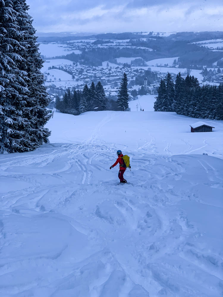 Melanie fährt mit ihrem Snowboard gekonnt über die Piste in Nesselwang. Im Hintergrund ist der Ort Nesselwang und weite Teile des Allgäus zu sehen. Erste Abfahrt 2021 zurück in Deutschland.