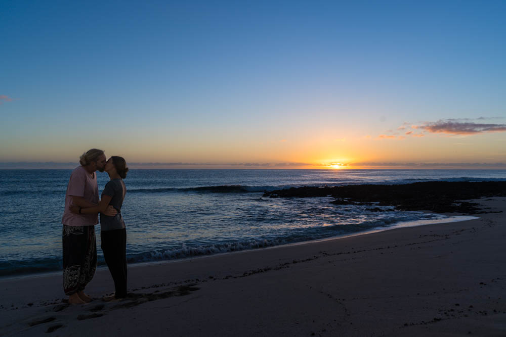 Melanie und Julian stehen küssend in den Dünen bei Corralejo und hinter ihnen geht über dem Atlantik die Sonne auf. Unsere Zeit auf Fuerteventura geht zu Ende