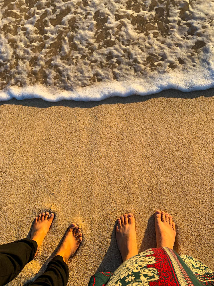 Melanie und Julian stehen im Sand in den Dünen bei Corralejo. Es sind die Beine und nackten Füße von den beiden zu sehen, sie stehen im Sand und das Wasser des Atlantiks wird als Welle zu ihnen geschwemmt. Unsere Zeit auf Fuerteventura geht zu Ende
