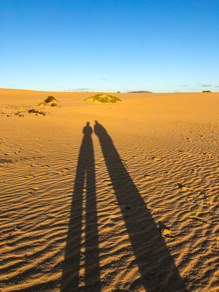 Es sind die Dünen bei Corralejo zu sehen sowie der strahlend blaue Himmel. Melanie und Julian werfen große Schatten in den Sand. Unsere Zeit auf Fuerteventura geht zu Ende