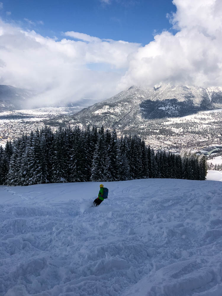 Julian bei der Abfahrt vom Hausberg in Garmisch. Er legt sich gekonnt in die Kurve und der Schnee spitzt ganz leicht nach oben. Im Hintergrund ist Garmisch und der Wank zu sehen.
