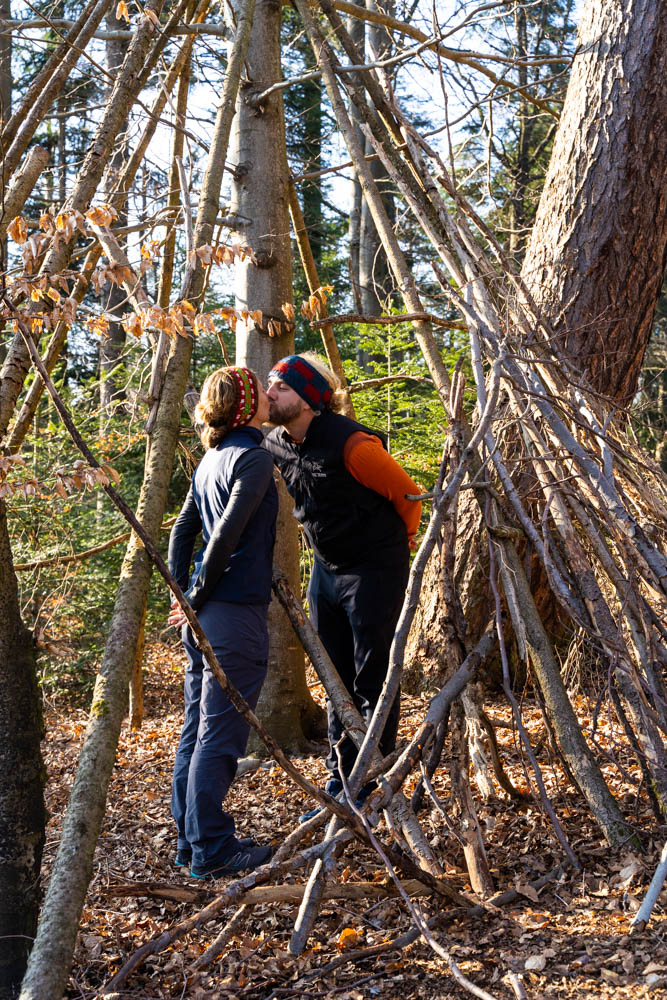 Melanie und Julian stehen in einem aus Ästen gebauten Unterstand im Wald und küssen sich. Es ist ihr erster längerer Spaziergang zurück in Deutschland.