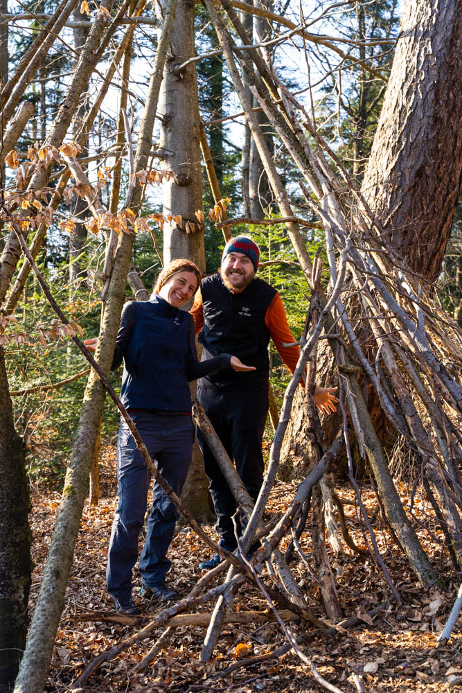 Melanie und Julian stehen in einem aus Ästen gebauten Unterstand im Wald und grinsen in die Kamera. Es ist ihr erster längerer Spaziergang zurück in Deutschland.