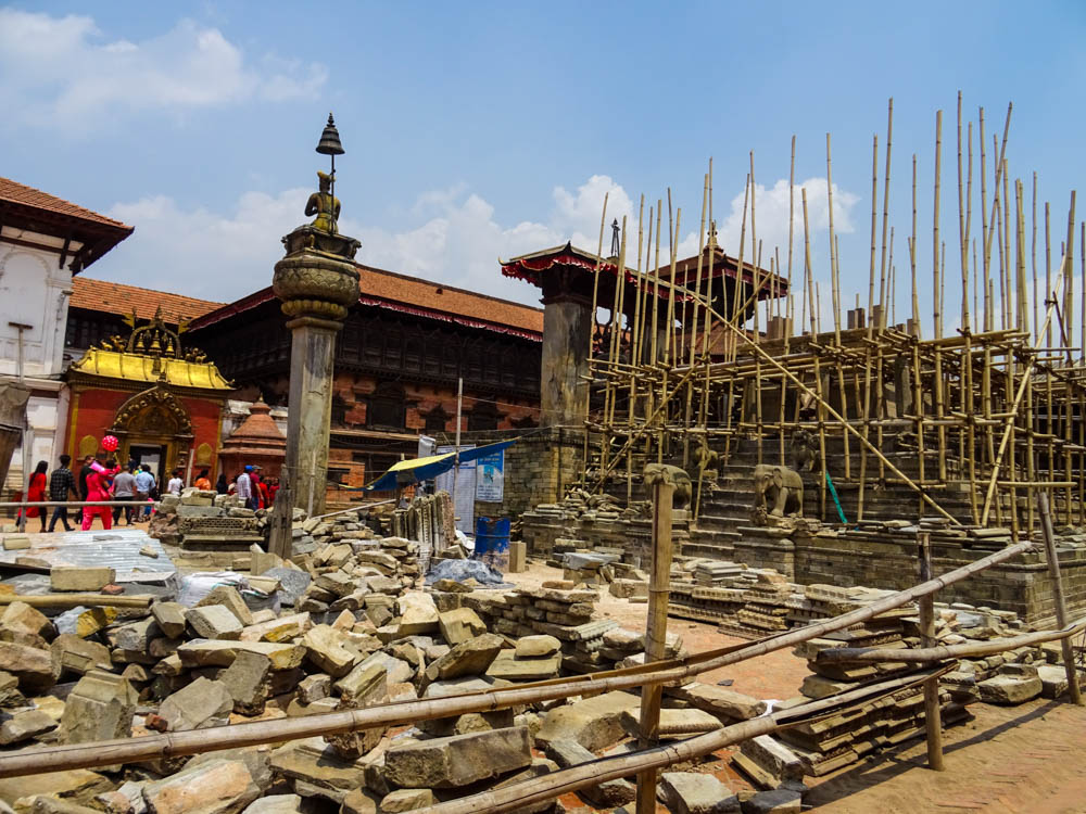 Auf einem Platz steht ein Gerüst aus Bambusstangen. Dahinter kann man den Sockel eines Tempels oder ähnlichem erkennen. Davor liegen Steine des ursprünglichen Tempels der beim Erdbeben zerstört wurde. Fernreise Nepal Eindrücke