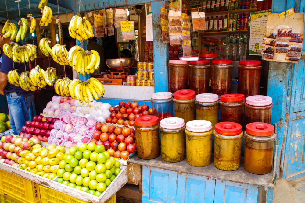 Straßenstand mit Lebensmittel in Bhaktapur. Es sind ein paar Glasfässer mit verschiedenen Inhalten zu sehen. Zudem liegen u.a. Äpfel unterschiedlicher Sorten aus und es hängen Bananen an Schnüren. Fernreise Nepal Eindrücke