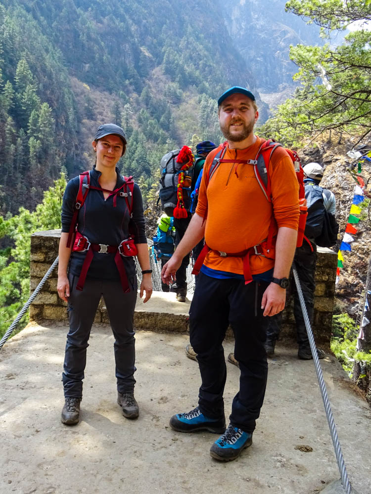 Melanie und Julian stehen vor der Edmund Hillary Bridge in Nepal beim Trekking zum Mount Everest Base Camp. Hinter ihnen staut es sich etwas, da hier viele Menschen drüber wandern. Es sind ein paar bunte Gebetsfahnen an der Brücke zu sehen.