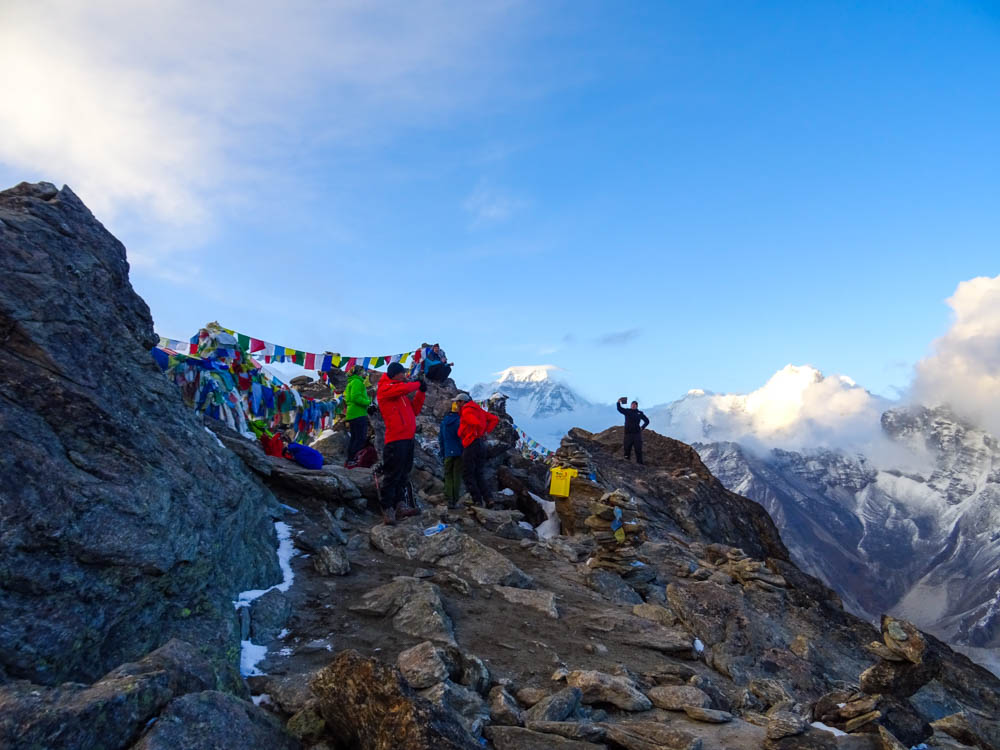 Bei dem Trekking zum Mount Everest Base Camp ging es auf den Gipfel Gokyo Ri. Es sind ein paar Wanderer und Julian auf dem Gipfelplateau. Bunte Gebetsfahnen sind an den Felsen befestigt. Der Himmel ist blau mit ein paar Wolken, im Hintergrund sind andere hohe Berge zu sehen.