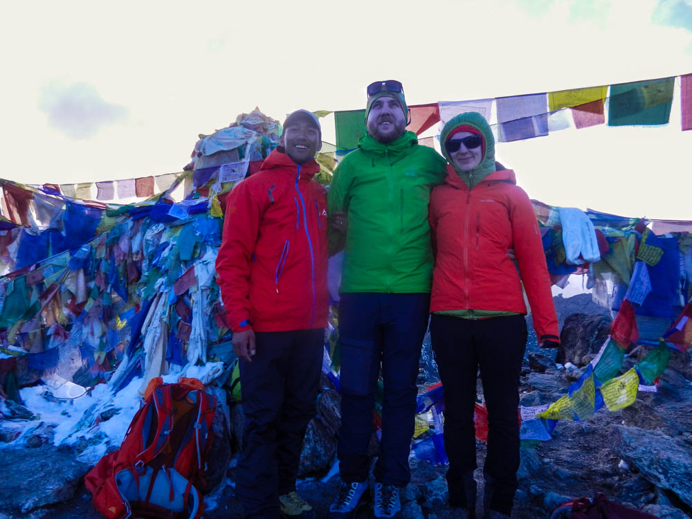 Melanie und Julian posieren mit Guide Keshar auf dem Gokyo Ri. Hinter ihnen sind etliche bunte Gebetsfahnen. Trekking Mount Everest Base Camp
