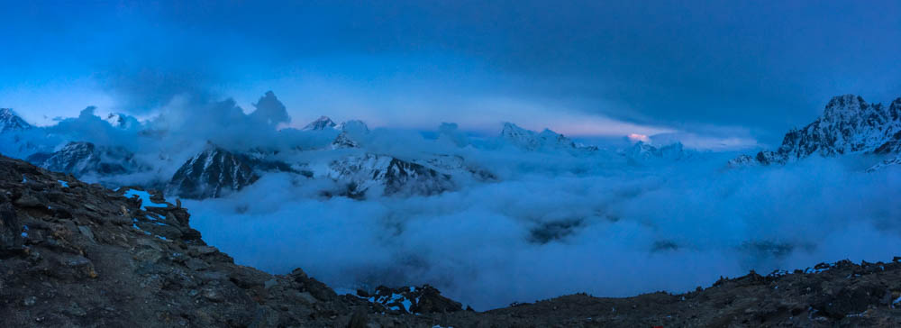 Blick vom Gokyo Ri auf das Himalaya Gebirge. Es ist Abend und der Himmel ist leicht orange-rot verfärbt. Im Tal und teilweise vor den Bergen hängen Wolken. Der Mount Everest ist mit einer roten Spitze zu sehen. Trekking Mount Everest Base Camp