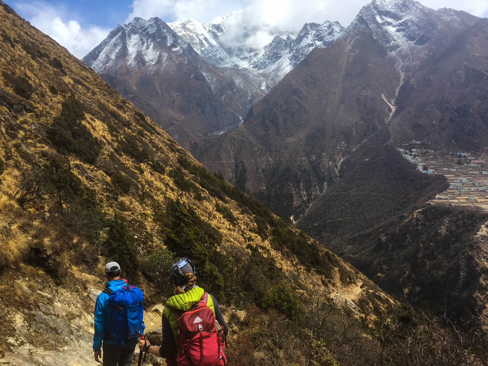 Melanie und Guide Keshar wandern gerade einen Pfad hinunter. Rechts in der Ferne ist ein Dorf auf einem Hügel zu erkennen, ansonsten sind einige Berge zu sehen. Trekking Nepal zum Mount Everest Base Camp.