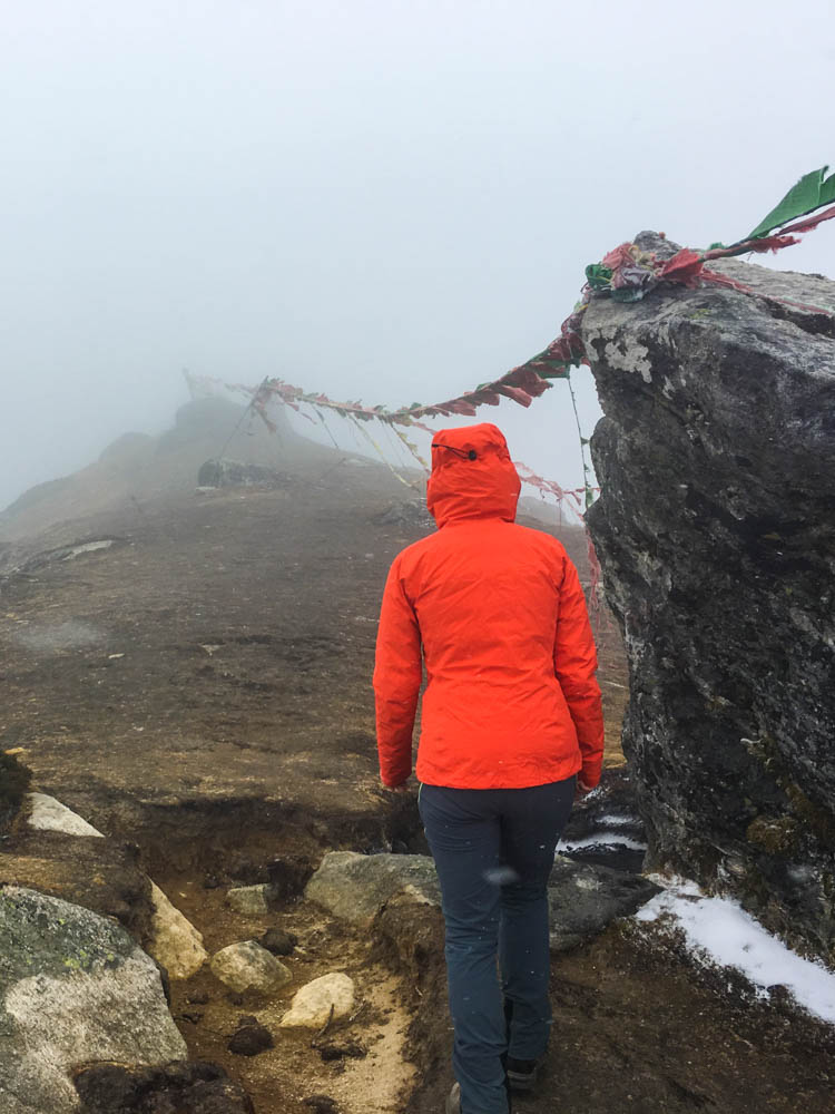 Akklimatisierungstour im Nebel auf dem Trekking zum Mount Everest Base Camp. Melanie läuft gerade an einem Felsen vorbei, an welchem bunte Gebetsfahnen angebracht sind. Sie bildet zum nebligen weiß ein bunter Kontrast mit ihrer orangenen Jacke.