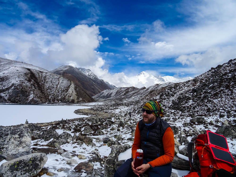 Julian macht Pause auf dem Trekking zum Mount Everest Base Camp. Er sitzt auf einem Stein und rund herum ist Schnee zu sehen. Julian bildet einen bunten Kontrast zu der weißen Umgebung, u.a. eingeschneiter See.