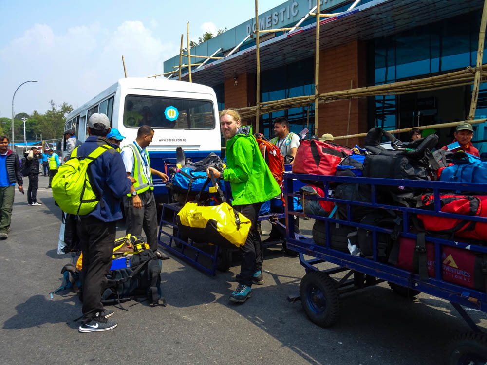Gepäck wird gerade am Flughafen in Kathmandu von einem Anhänger abgeladen. Julian hat gerade unsere Expeditionstasche in der Hand. Julian hat sich als Porter ausprobiert. Er hat gerade noch unser komplettes Gepäck mit einem Band an der Stirn befestigt, unser Porter hilft ihm dabei es abzunehmen.