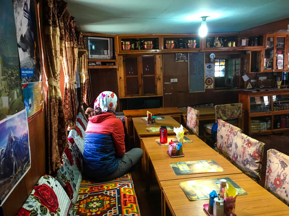 Melanie sitzt in einer Lodge in Nepal und schaut Richtung Küche. Überall hängen Bilder an der Wand. Über dem Tresen steht sogar ein kleiner Röhrenfernseher.