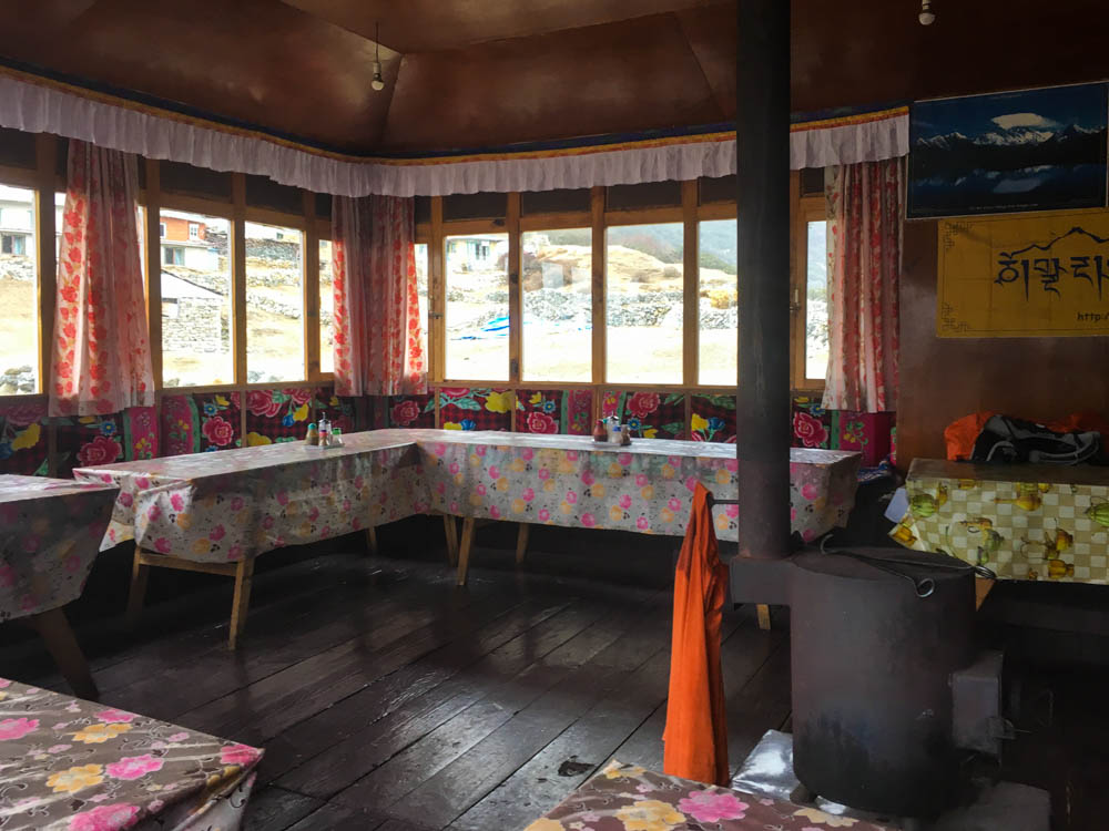 Aufnahme von einem klassischen Speiseraum in einer Lodge in Nepal. Es gibt viel Fensterfläche um nach draußen zu schauen. Es sind bunte Tischdecken auf den niedrigen Tischen. In der Mitte des Raumes steht ein Ofen.