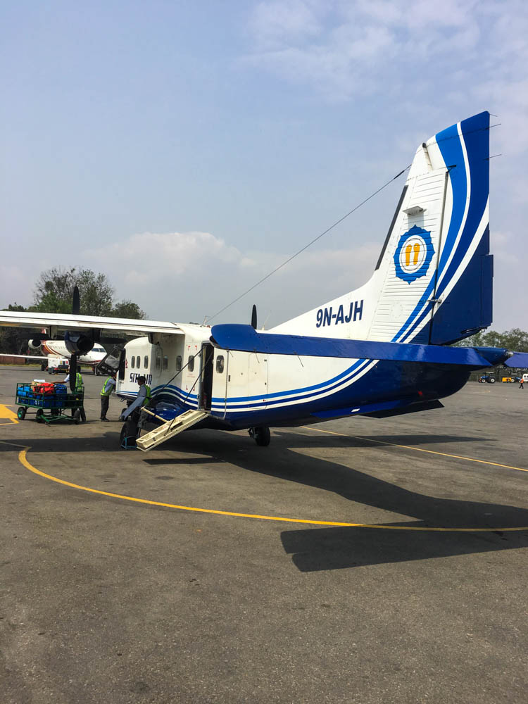 Eines der kleinen Flugzuege die nach Lukla fliegen steht in Kathmandu auf dem Rollfeld. Im hinteren Teil ist die Einstiegstüre ausgeklappt. Am Cockpit steht ein Handkarren mit Gepäck, das gerade in die Nase des Flugzeugs eingeladen wird.