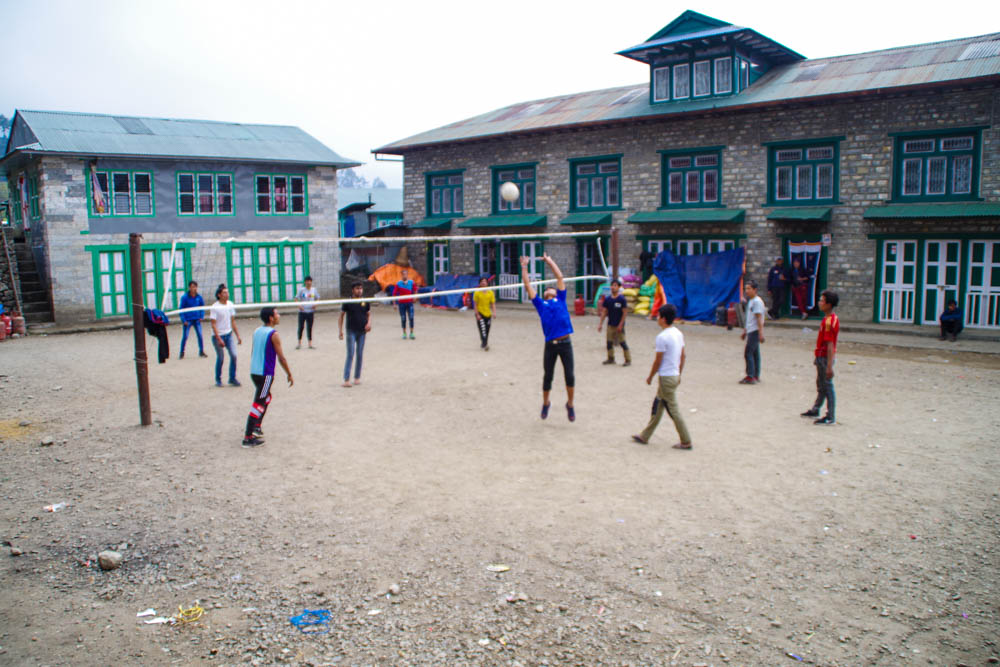 Auf einem Feld mitten im Dorf Lukla spielen junge Menschen Volleyball. Drum herum stehen zwei Häuser.