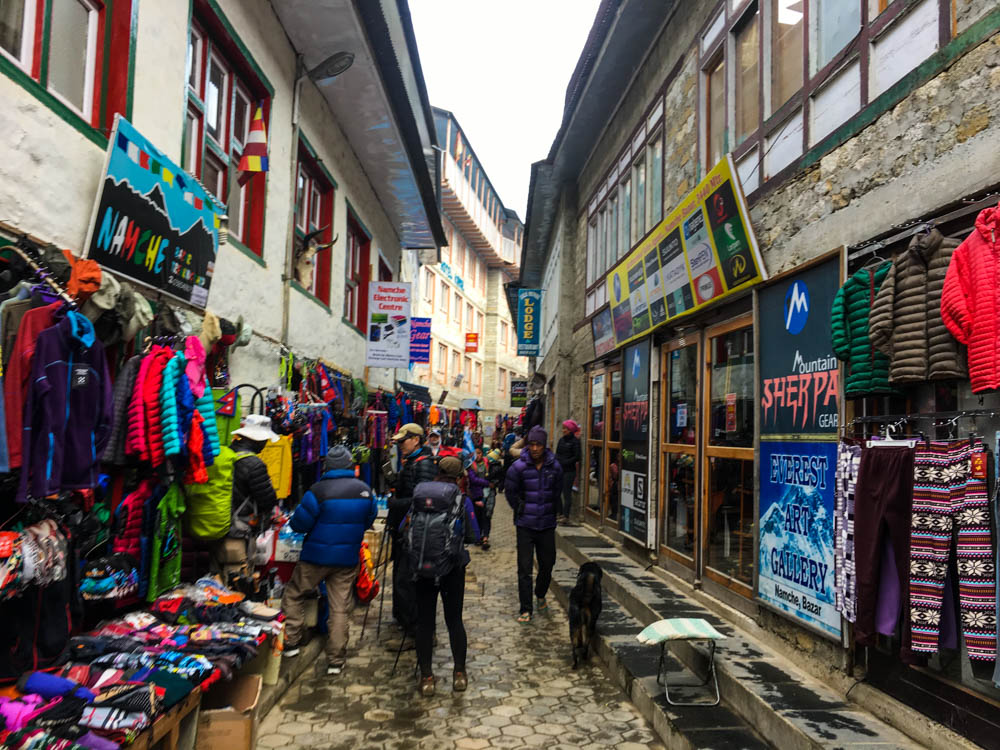Einkaufsviertel in Namche Bazar. Vor den Läden steht Outdoor-Kleidung und durch die Gasse laufen mehrere Touristen und Einheimischen. Alle bereit für ein Trekking in Nepal