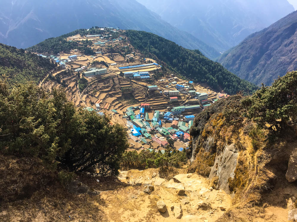 Vogelperspektive auf Namche Bazar. Dorf liegt u- und terrassenförmig im Hang. Die bunten Dächer der Häuser stechen ins Auge, drum herum sind grüne Büsche und gegenüber ein weiteres Bergmassiv. Nepal Trekking zum Mount Everest Base Camp.