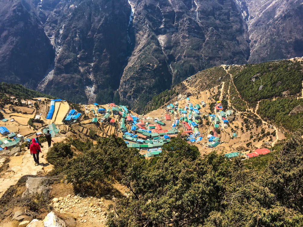 Vogelperspektive auf Namche Bazar. Dorf liegt u- und terrassenförmig im Hang. Die bunten Dächer der Häuser stechen ins Auge, drum herum sind grüne Büsche und gegenüber ein weiteres Bergmassiv. Nepal Trekking zum Mount Everest Base Camp.