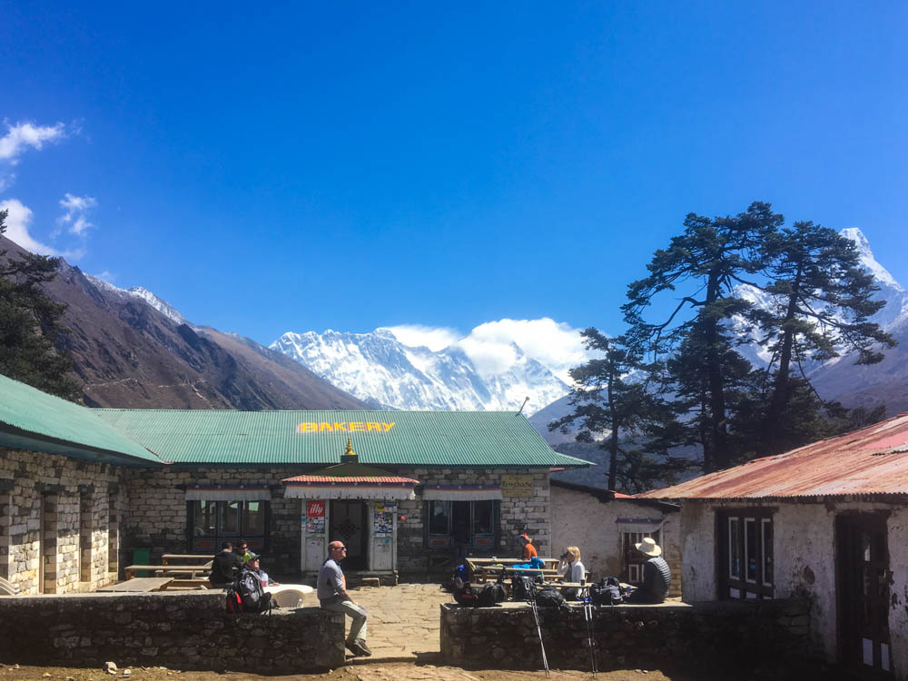Es ist eine Bäckerei in Techboche zu sehen. Vor der Bäckerei sitzen ein paar Menschen, dahinter ist der Mount Everest zu sehen. Der Himmel ist strahlend blau. Trekking Fortsetzung