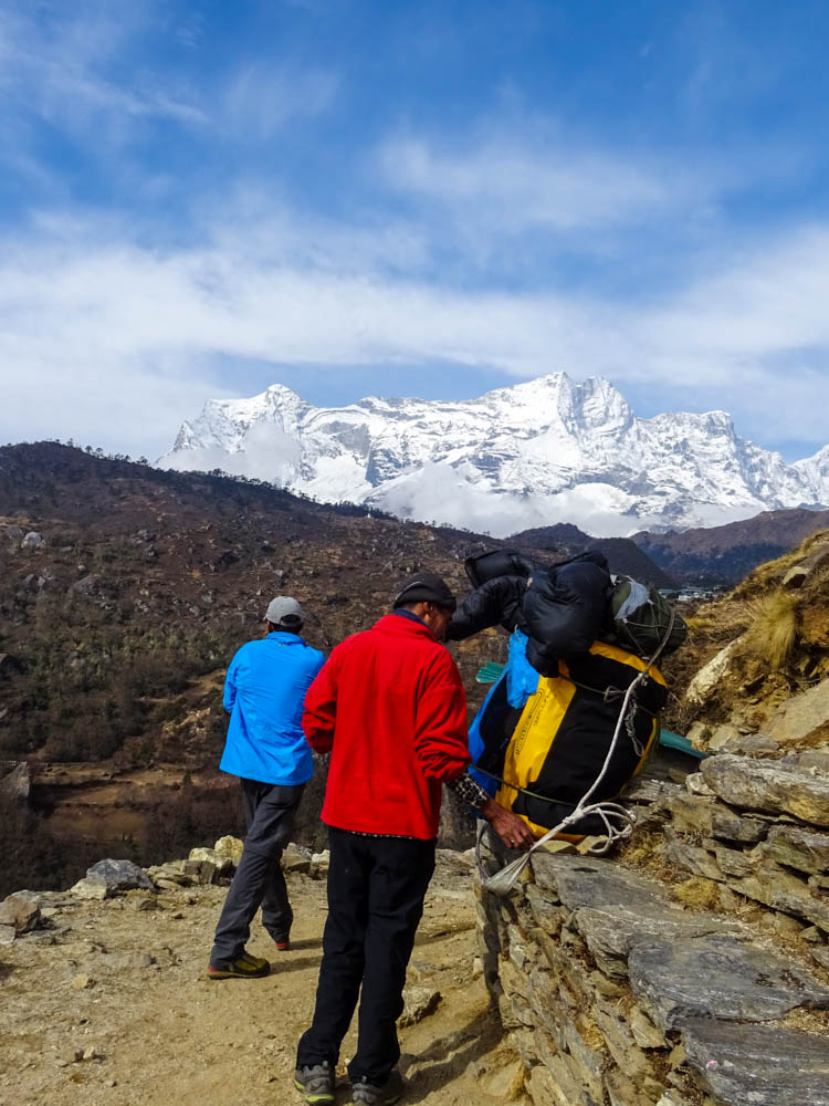 Pause auf dem Trekking in Nepal. Es sind Guide und Träger mit unserem Gepäck zu sehen. In der Ferne sind weißbedeckte Gipfel zu sehen, der Himmel ist blau und leicht bewölkt.