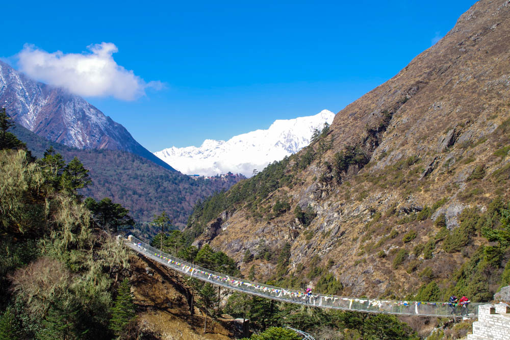 Hängebrücke in Nepal. Links Wald, rechts Berg. Im Hintergrund sind schneebedeckte Berge zu sehen, der Himmel ist nahezu strahlend blau. Trekking Mount Everest Region Fortsetzung