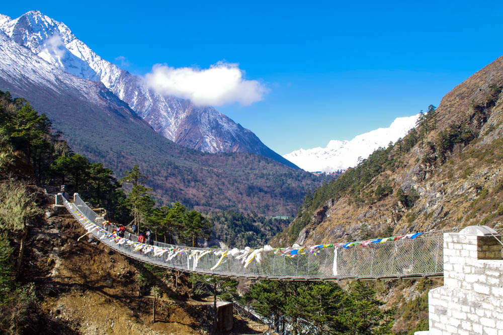 Hängebrücke in Nepal. Links Wald, rechts Berg. Im Hintergrund sind schneebedeckte Berge zu sehen, der Himmel ist nahezu strahlend blau. Trekking Mount Everest Region Fortsetzung