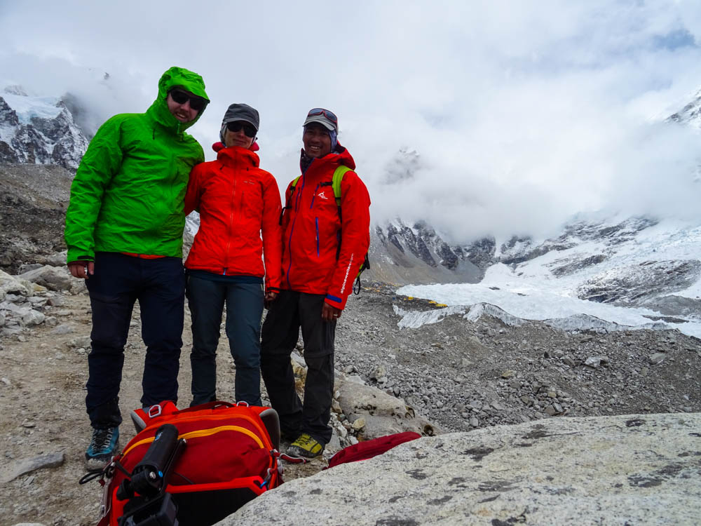 Melanie, Julian und Guide Keshar stehen vor dem Mount Everest Base Camp. Hinter ihnen ist der Khumbu-Gletscher zu sehen, Wolken hängen tief und ziehen immer mehr rein. Trekking Fortsetzung