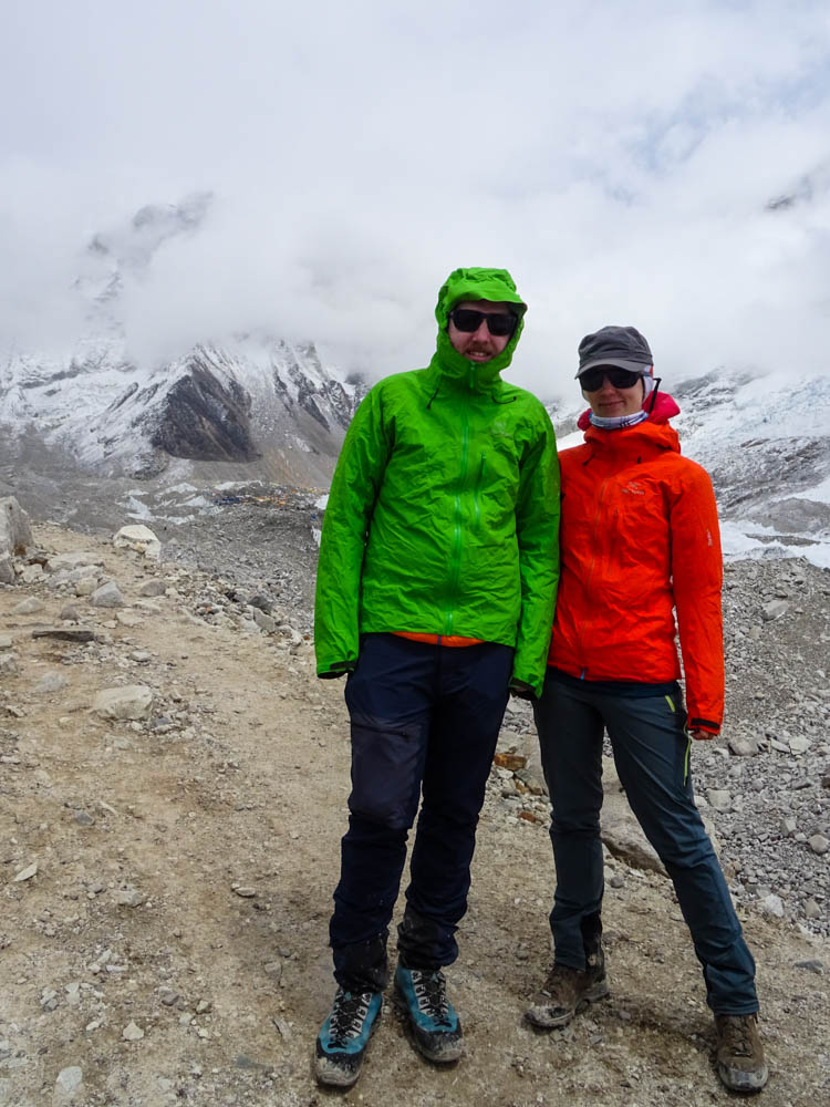 Melanie und Julian posieren vor dem Mount Everest Base Camp. Die Wolken hängen tief. Das Outfit der beiden sticht dank den kräftigen Farben grün und orange heraus.