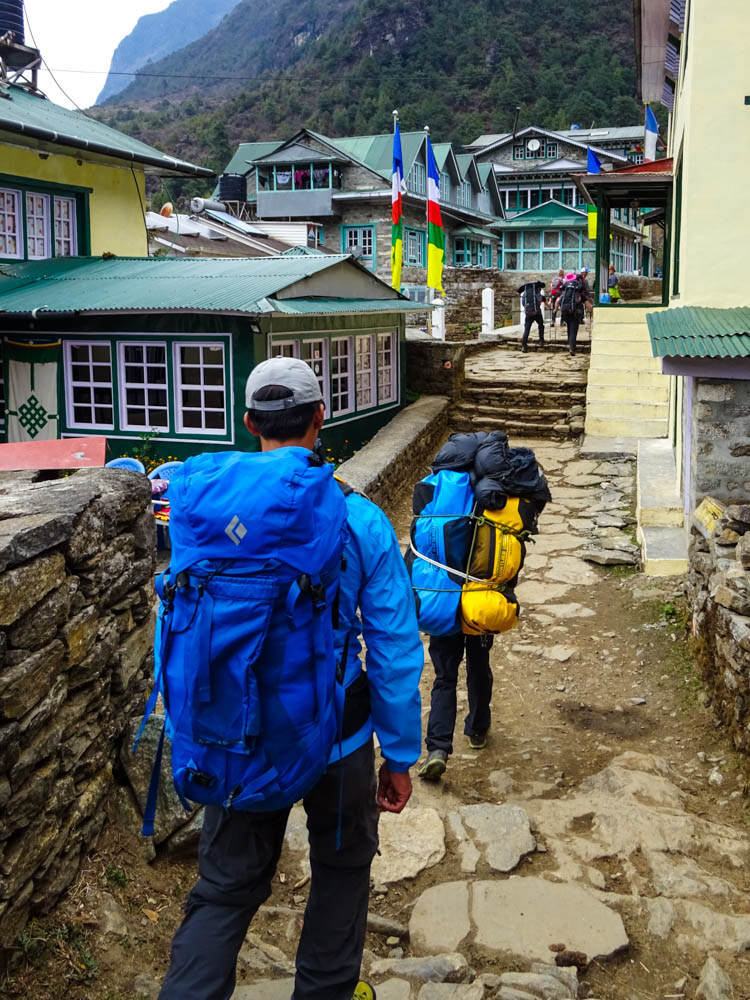 Unser Guide Keshar und Träger Balkumar laufen durch eine schmale Gasse in einem Dorf. Es sind typische Lodges zu sehen. Im Hintergrund sieht man zwei Fahnen mit den typischen Farben blau, weiß, rot und grün wehen.