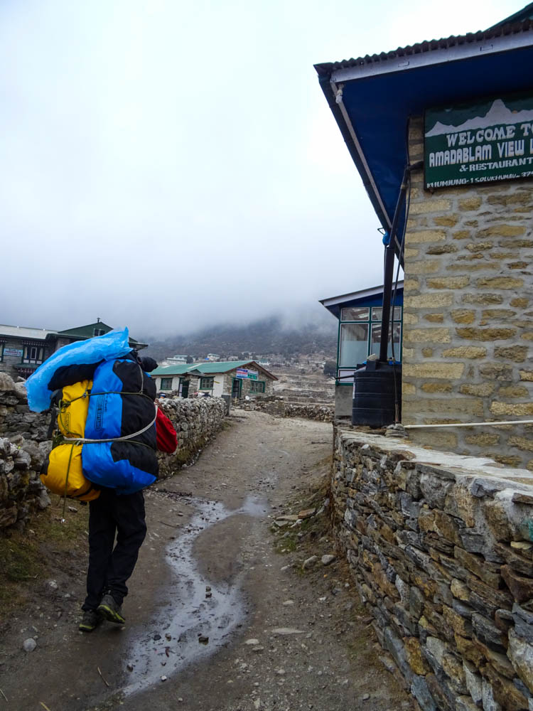 Porter beim Trekking in Nepal zum Mount Everest Base Camp. Dieser trägt unser Gepäck und läuft gerade durch ein Dorf. Der Himmel ist komplett bewölkt.