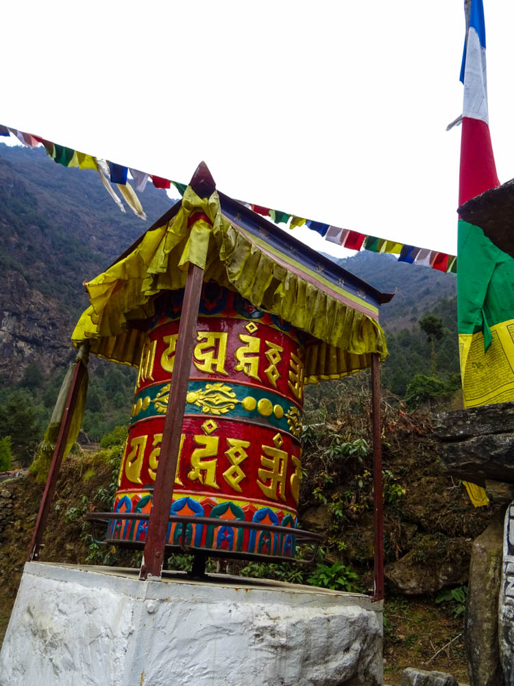 Es ist eine bunte Gebetsmühle sowie bunte Gebetsfahnen zu sehen. Diese stehen am Wegesrand für ein Trekking zum Mount Everest Base Camp.
