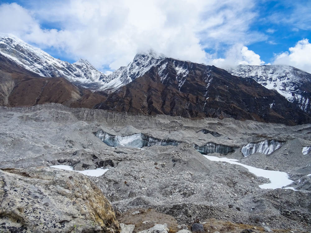 Ngozumpa Gletscher mit dahinter liegenden Bergen. Es ist viel Schutt zu sehen, aber auch Eisflächen innerhalb des Gletschers.